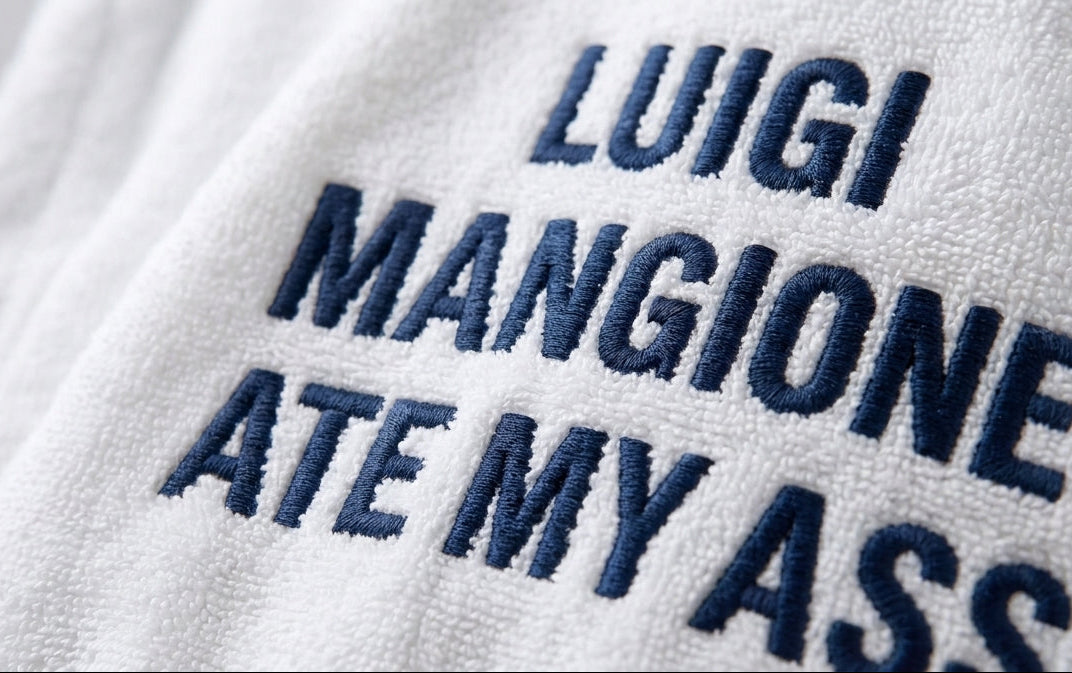 A close-up of a white towel with dark blue embroidered text reading 'LUIGI MANGIONE ATE MY ASS', shown against a softly blurred bathroom background.